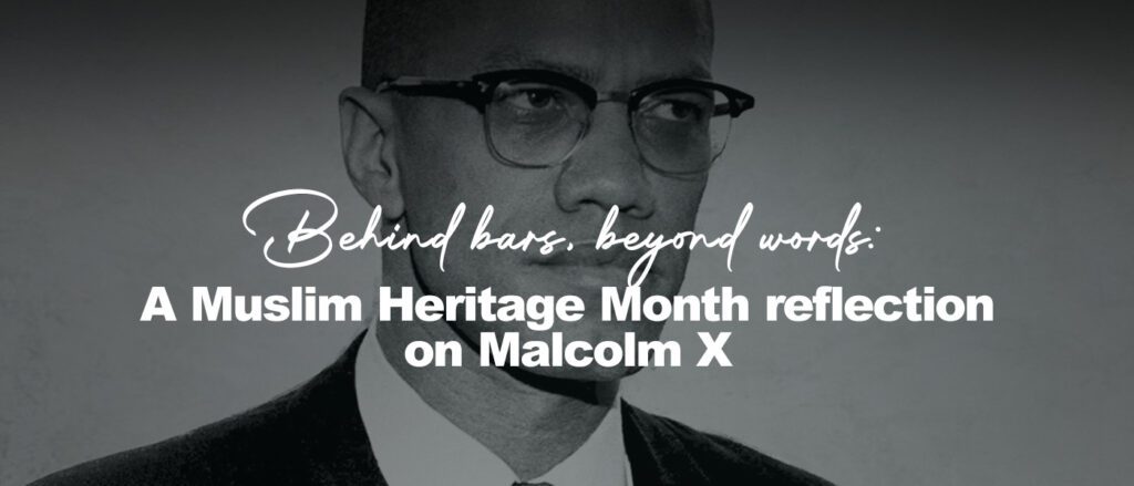 Black-and-white portrait of Malcolm X, shown from the chest up, wearing a dark suit, white shirt, and tie. He looks slightly to the right with a calm, serious expression, his round glasses catching soft light against a textured, light-colored background.